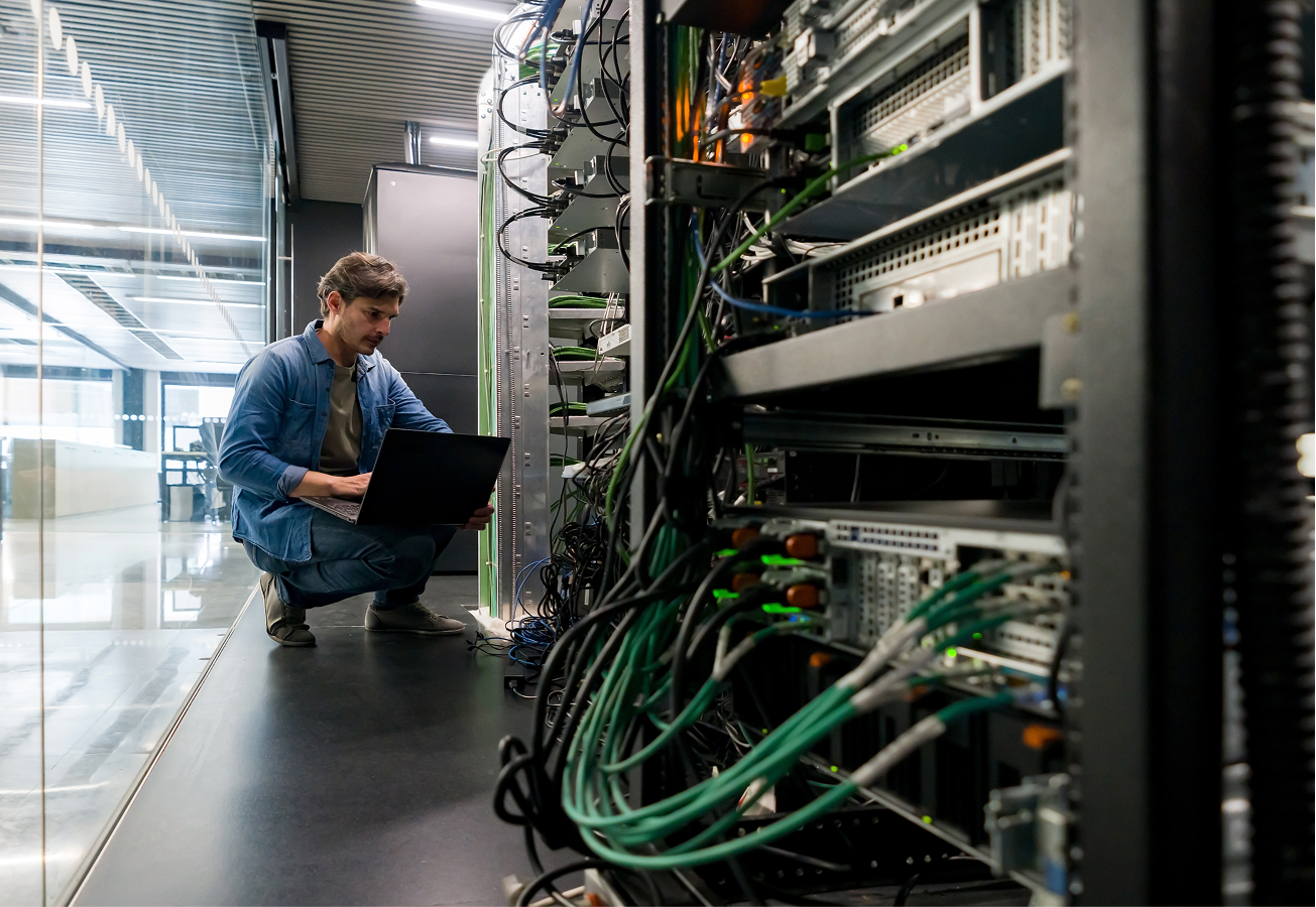 A man kneeling with a laptop reviewing wires in a server room.